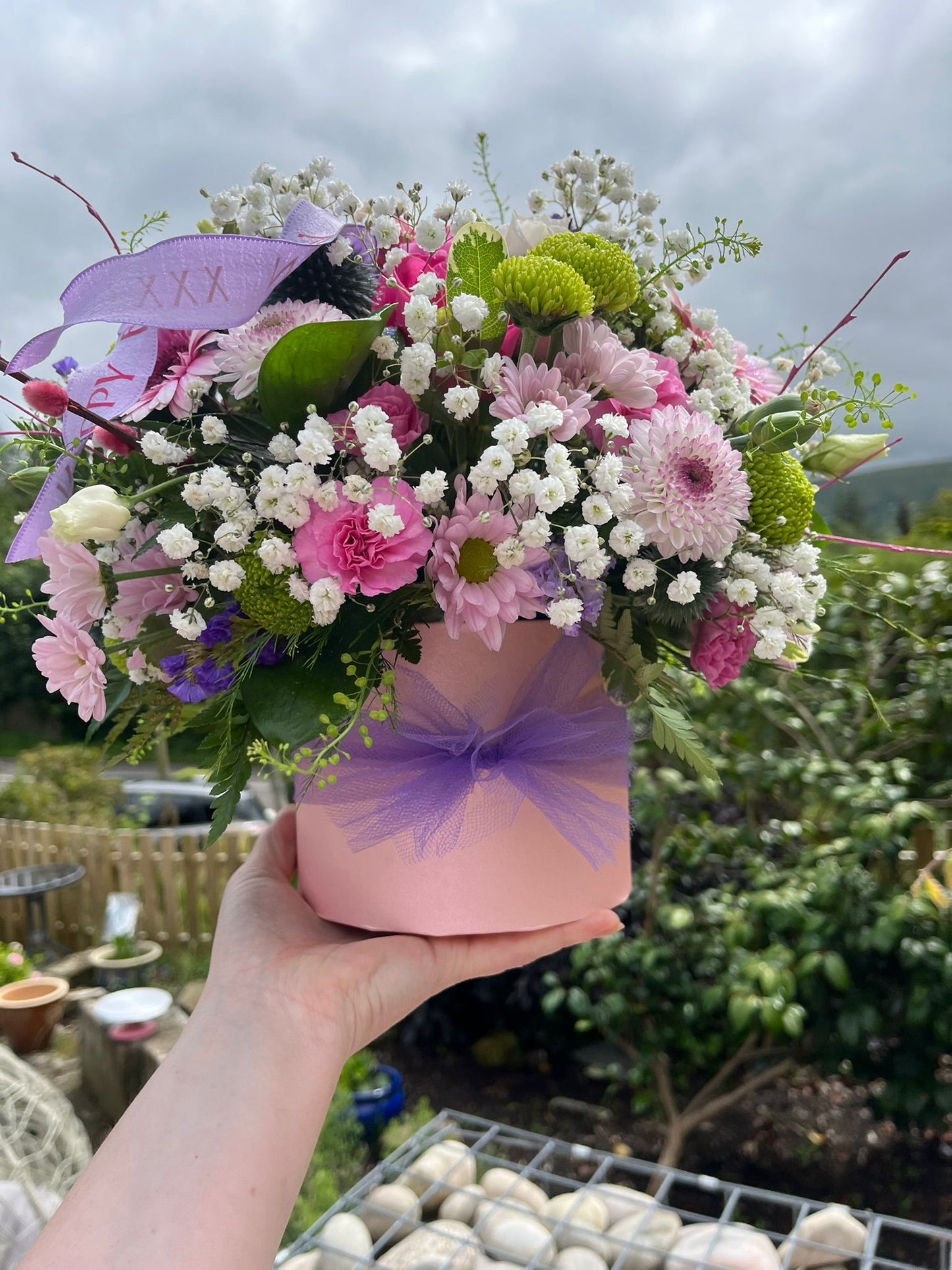 Bouquet of flowers in a pink box with a purple bow held by a person outdoors.