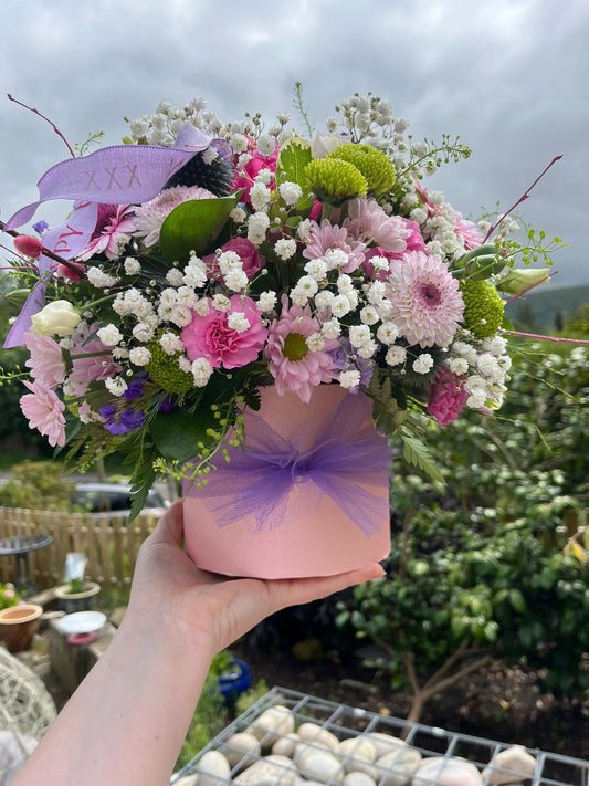 Bouquet of flowers in a pink box with a purple bow held by a person outdoors.